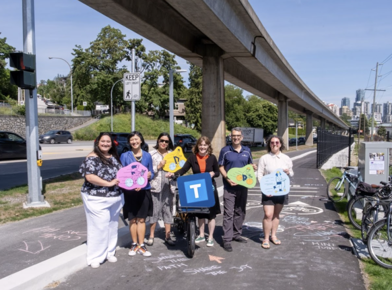 A group of seven people stand smiling with colorful signs shaped like bicycle silhouettes under an elevated SkyTrain track. They are gathered on a multi-use path marked with chalk messages and a "Welcome to BC Parkway" sign.