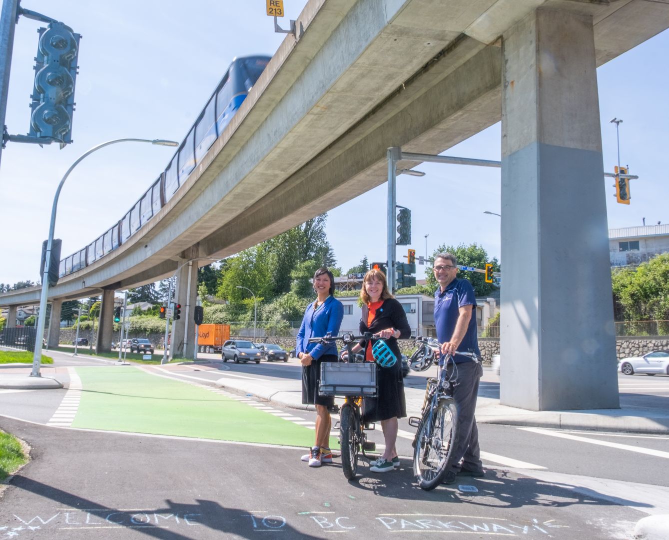 Three people with bicycles stand smiling on a multi-use path beneath an elevated SkyTrain track. They are surrounded by a green bike lane, traffic signals, and a concrete overpass. Chalk text on the path reads, "Welcome to BC Parkway."