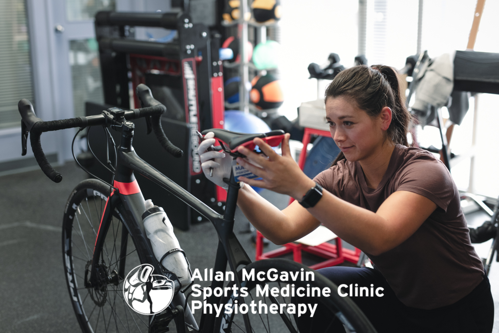 A Woman adjusts the seat of a road bike with text in white Allan McGavin Sports Medicine Clinic overtop of the image with a logo.