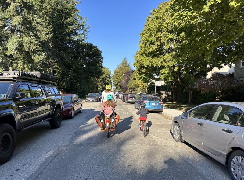 A mother and her two children ride their bikes on a residential roadway in Vancouver. Cars are parked on both sides of the street.