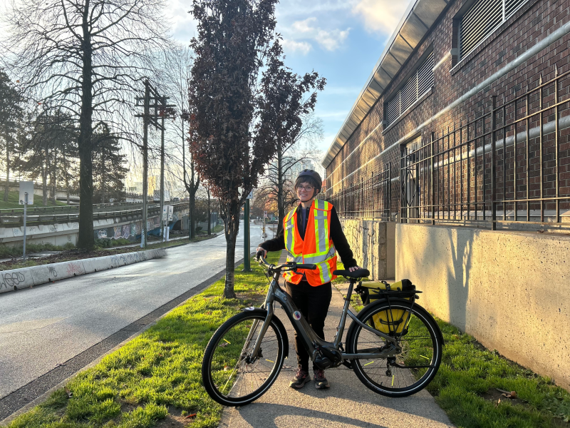 Chelsea standing on the sidewalk with an orange reflective vest and with the ohm e-bike.