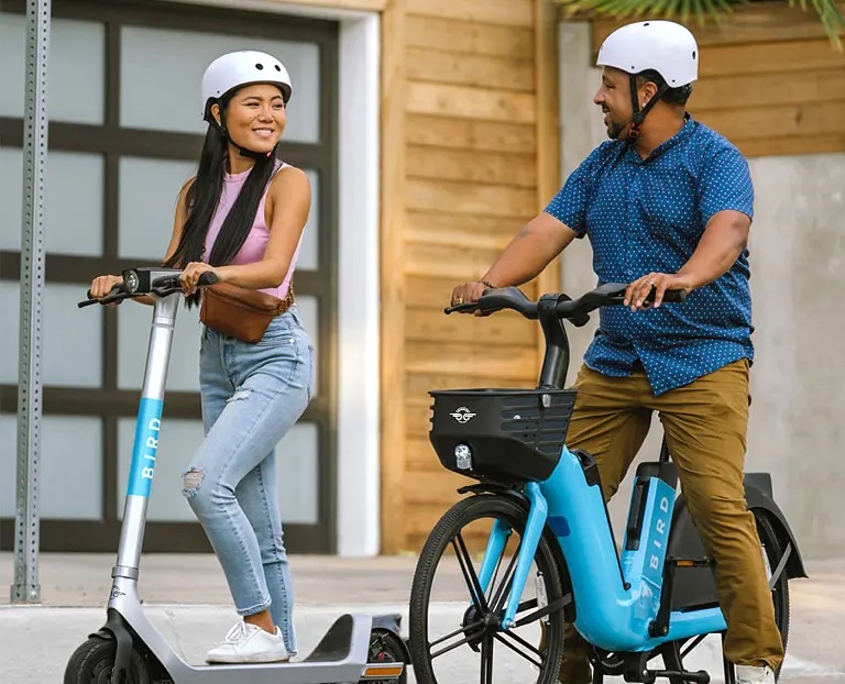 A Female on a bird scooter (left) wearing a white helmet and a man in a white helmet rides beside (right) on a blue and back Bird branded bike