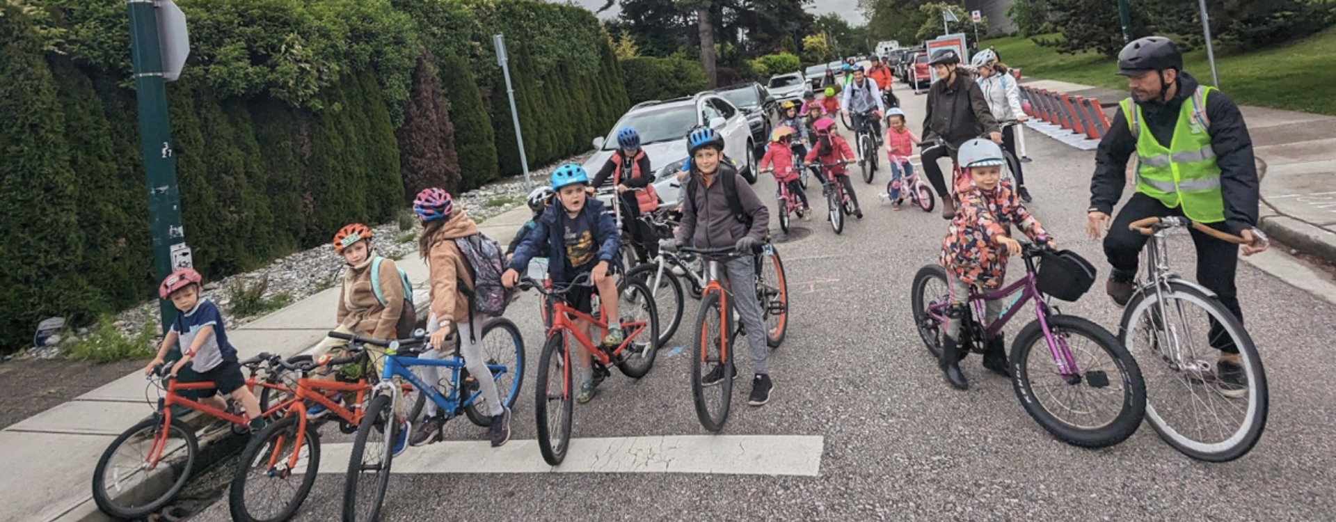 A group of about twenty students led by a couple of adults ride their bikes on a residential street as part of a "bike bus".