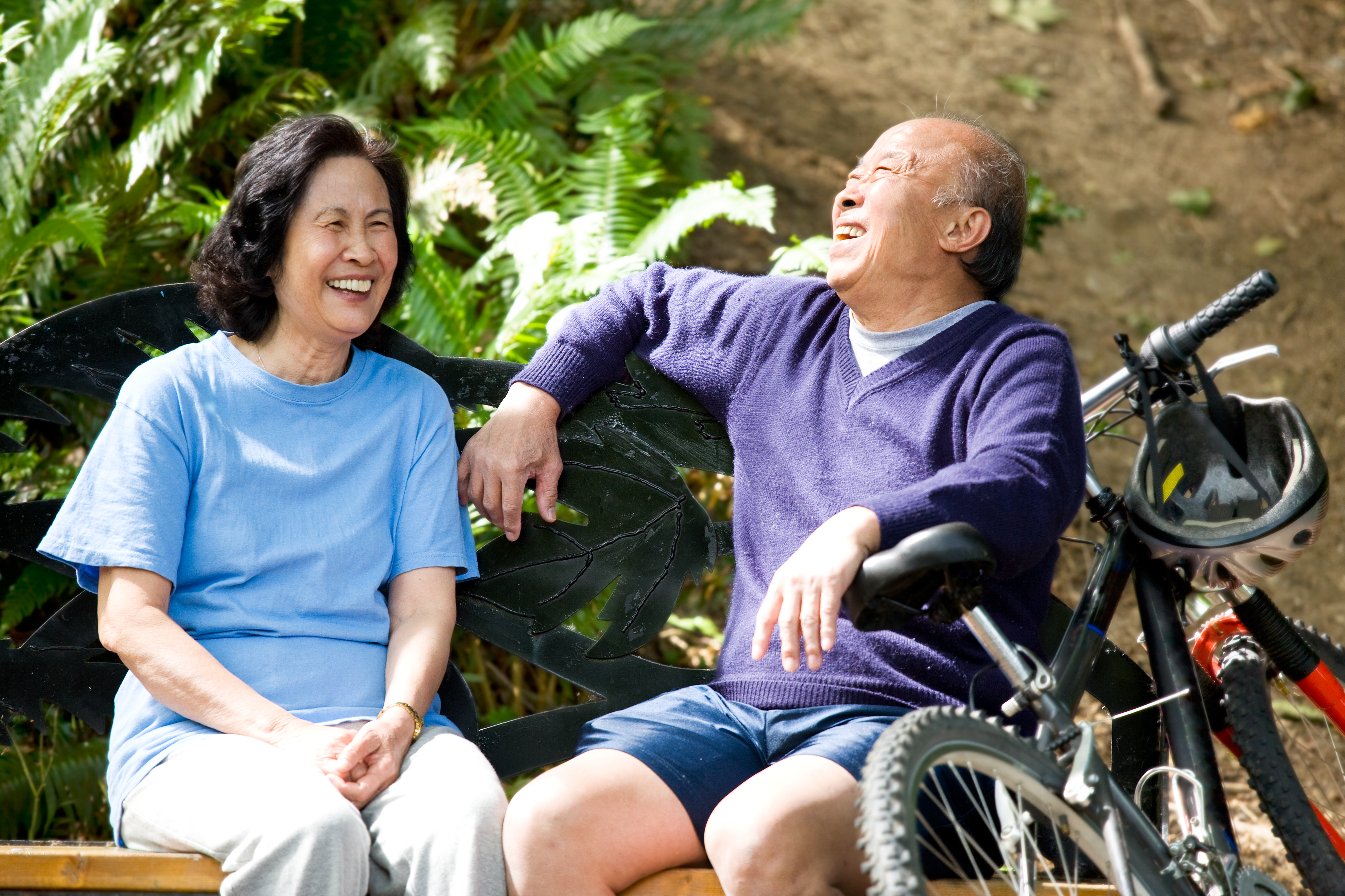 A senior couple laughs together while sitting on a bench. There is a bike beside the man.