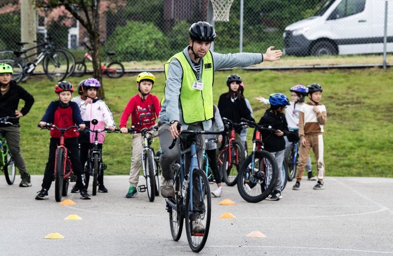 A bike education instructor shows how to make a left-hand turn signal. Several students on bikes watch.