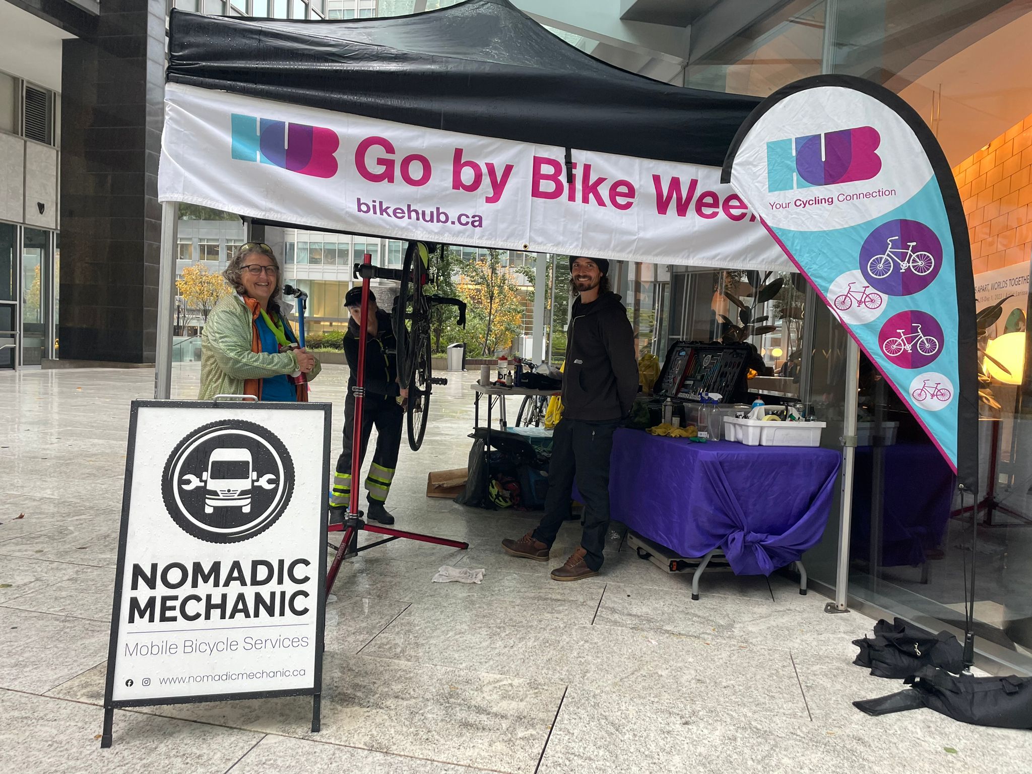Two male bike mechanics from Nomadic Mechanic and a female volunteer stand at a Go by Bike Week Celebration Station.