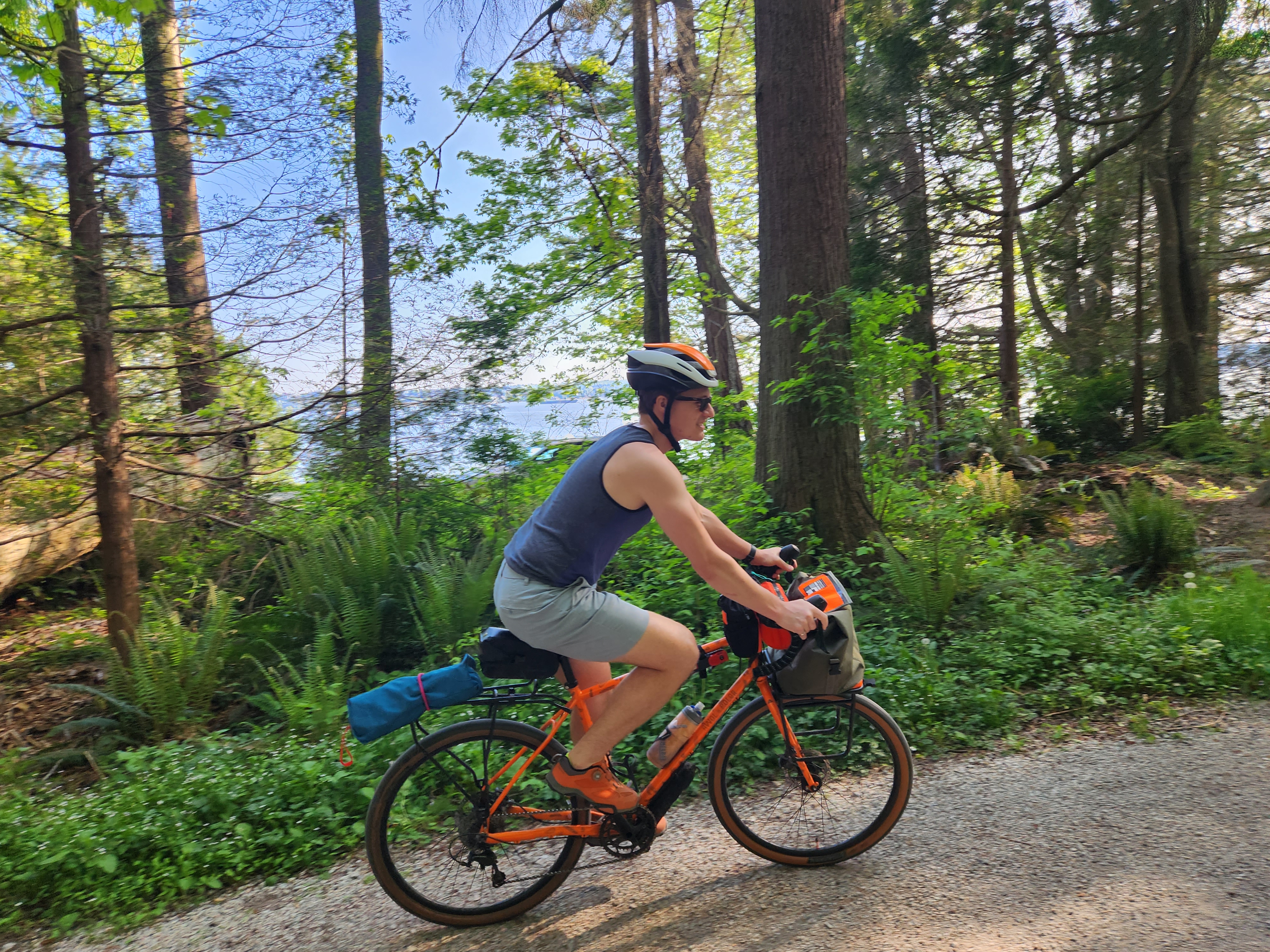 A man in his 30s rides his bike down a gravel trail. We see a peak-a-boo view of the ocean through the surrounding trees.