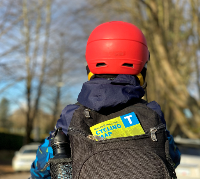 A boy wearing a helmet rides a bike. He is wearing a backpack with a cycling map sticking out of it.