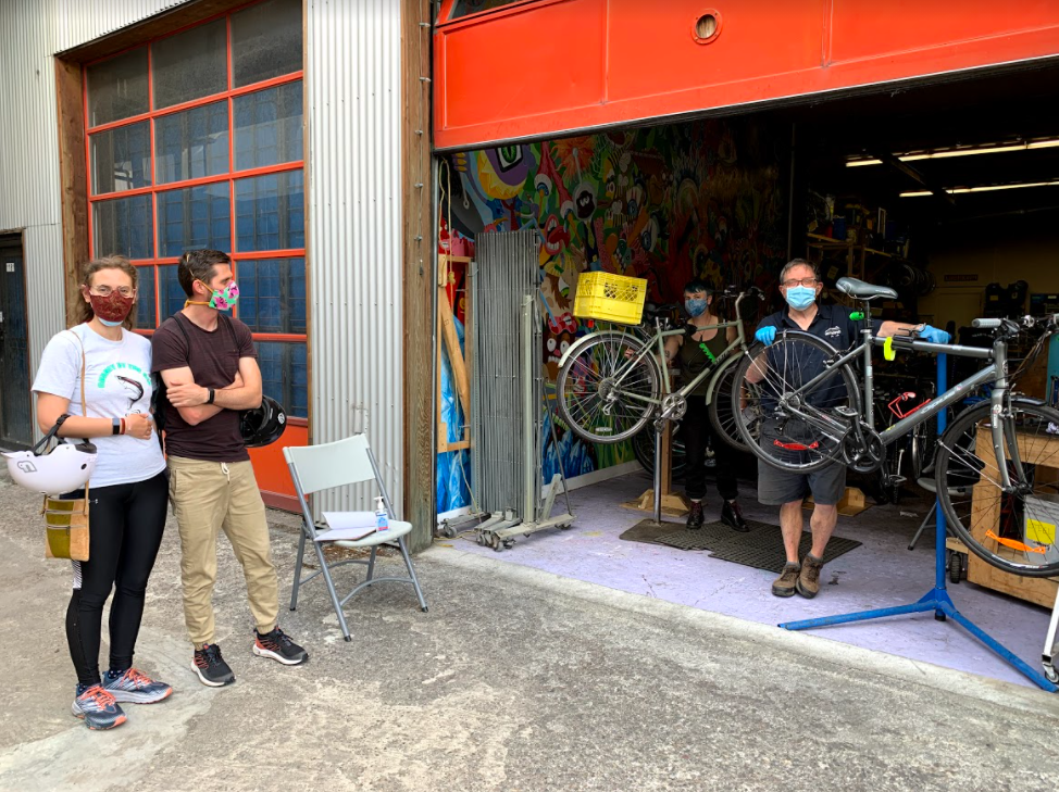 A man and woman stand wearing masks as they wait for their bikes to be repaired by two bike mechanics
