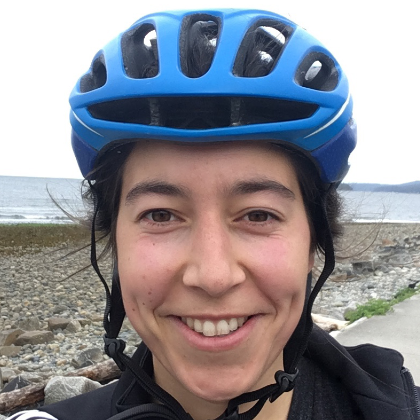 A portrait picture of Giovanna Lanius-Pascuzzi. She is a young white woman in her mid-20s. She has black hair tied back and wears a blue bike helmet. She smiles for the camera.