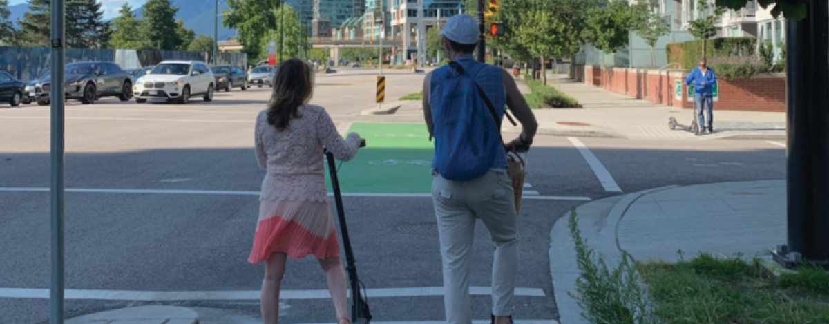A young woman and man in their 20s are stopped at an intersection in Vancouver. They are riding e-scooters. Their backs are to the camera.