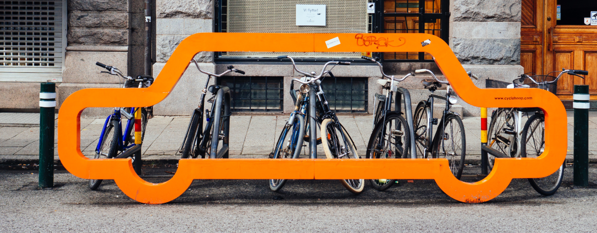 A row of five bike racks with seven bikes locked to them. The row of bike racks is on the side of the road. A metal installation that shows the outline of a car is in front of the row of bike racks.