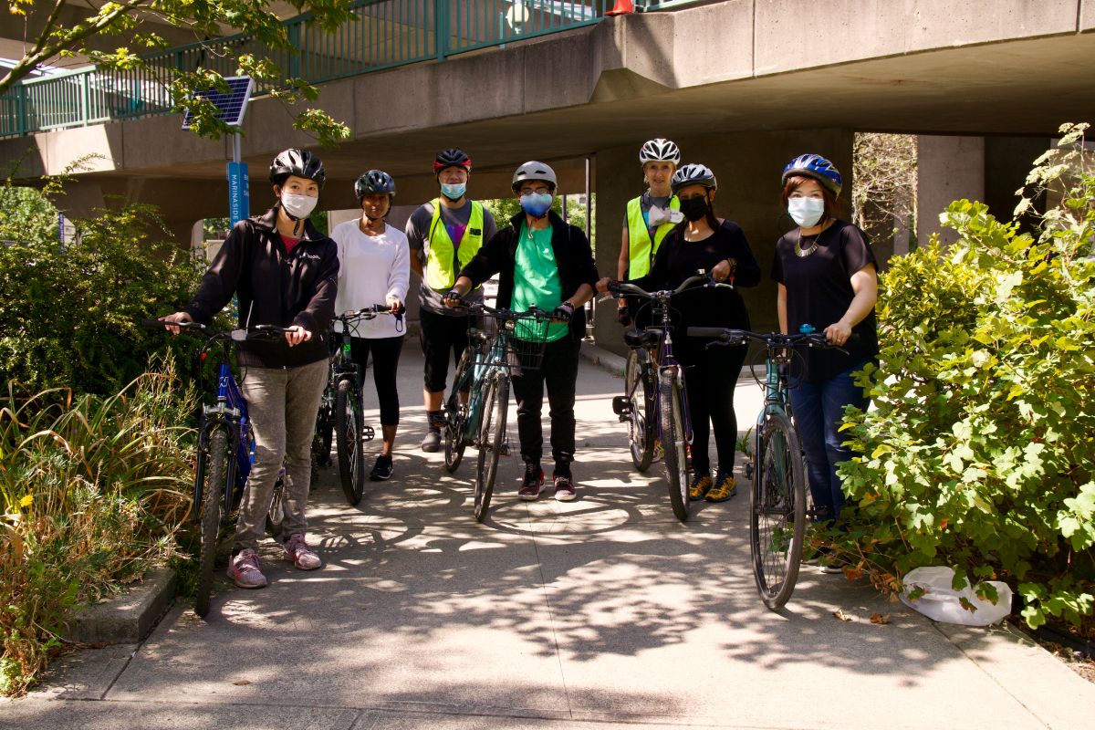 A group of four students and two bike education instructors stand in front of an overpass with their bikes