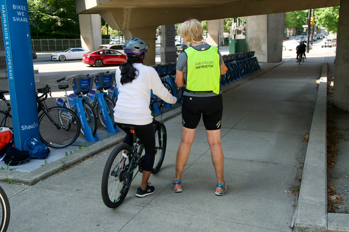 A female-presenting bike education instructor talks to a a student cycling. They are beside a Mobi by Shaw Go bike share station.