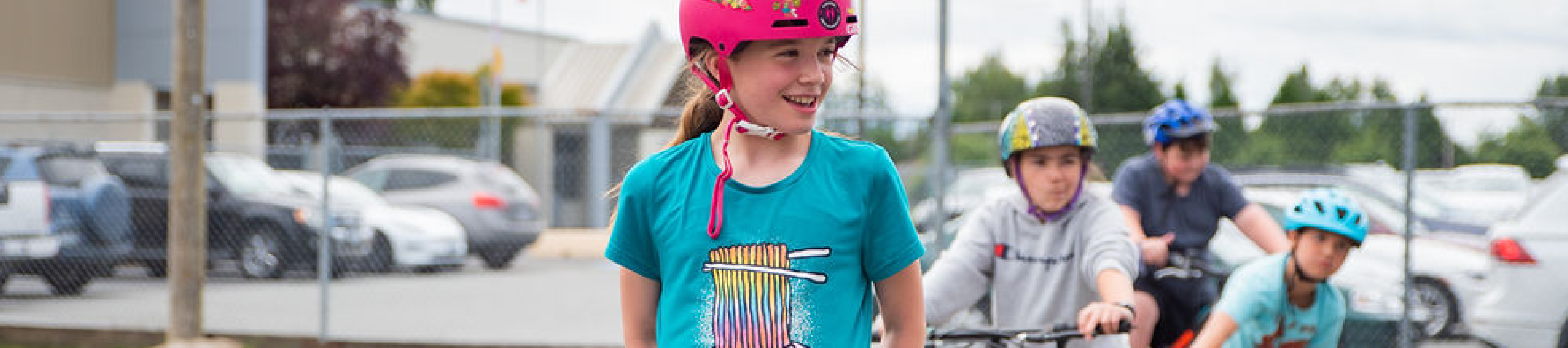 A young girl about 11 years old sites on her bike and smiles off into the distance. She is wearing a blue shirt and pink helmet. 