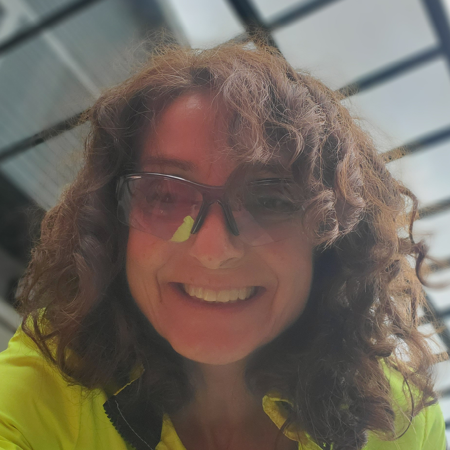 A photo of Esther Holobuwich - a woman in her 50s with short, curly brown hair. She is wearing clear cycling glasses and smiling.