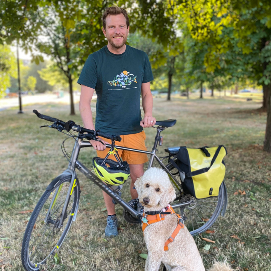 A photo of Luke Gillies - a man in his 40s with short brown hair. He is standing with his bike a doodle dog.