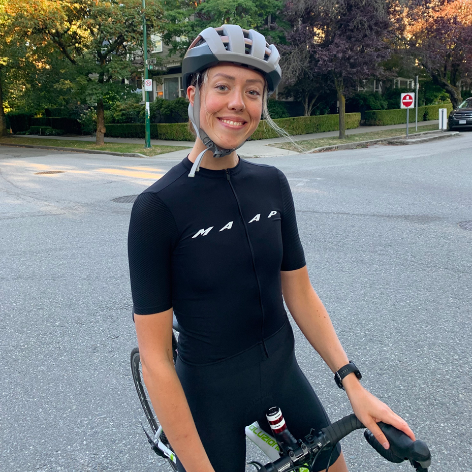 A photo of Mairin Shields-Brown - a woman in her late 20s/early 30s with blonde hair and blue eyes. She is wearing a helmet and standing with her bike on the side of a road.