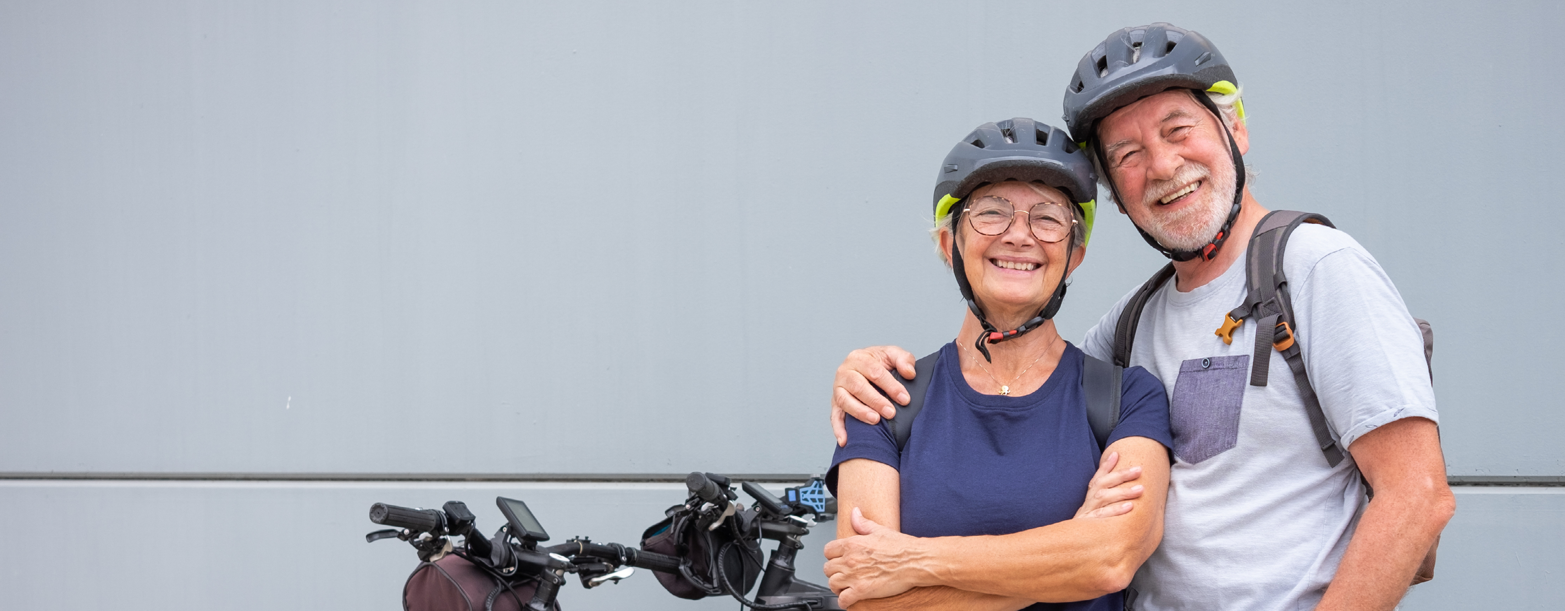 A white couple in their 60s stand beside their two e-bikes and smile for the camera. They are both wearing black helmets.