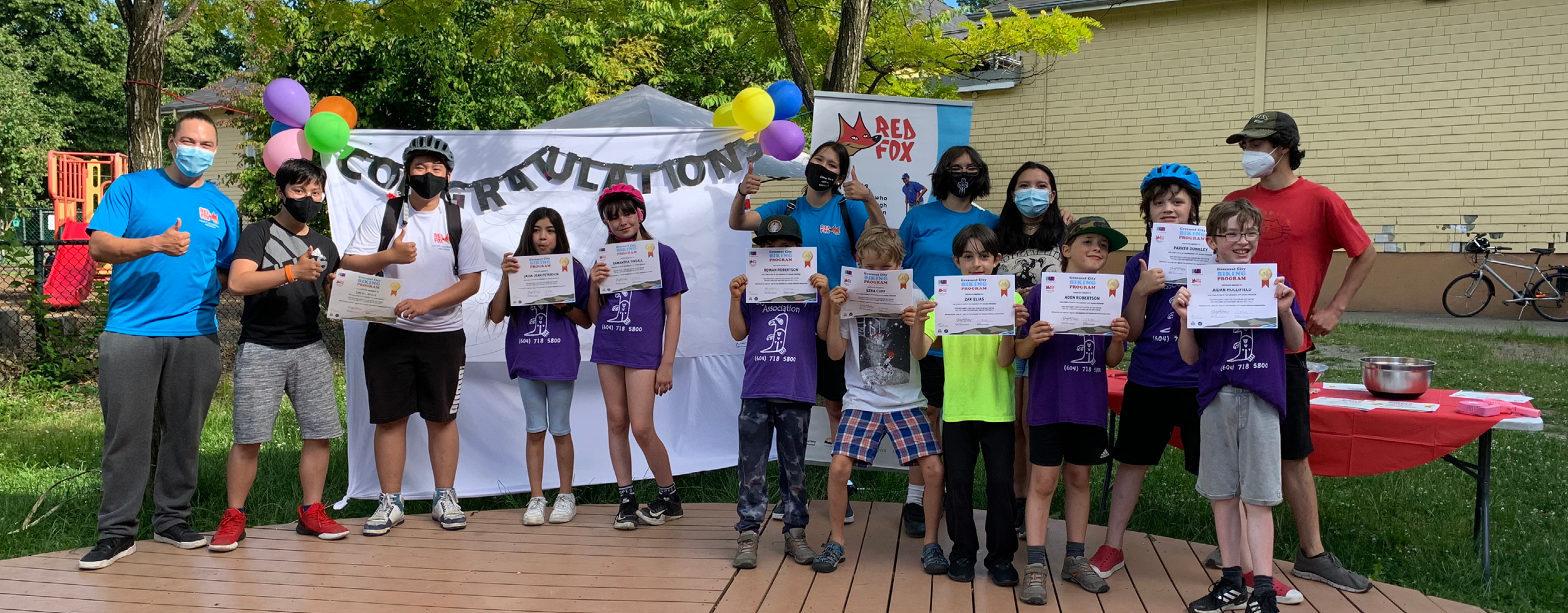 A group of young elementary school-aged students and four instructors stand in a row. The students are holding up their certificates of completion for completing the Ride Your Path course with Red Fox. This photo was taken at the graduation ceremony.