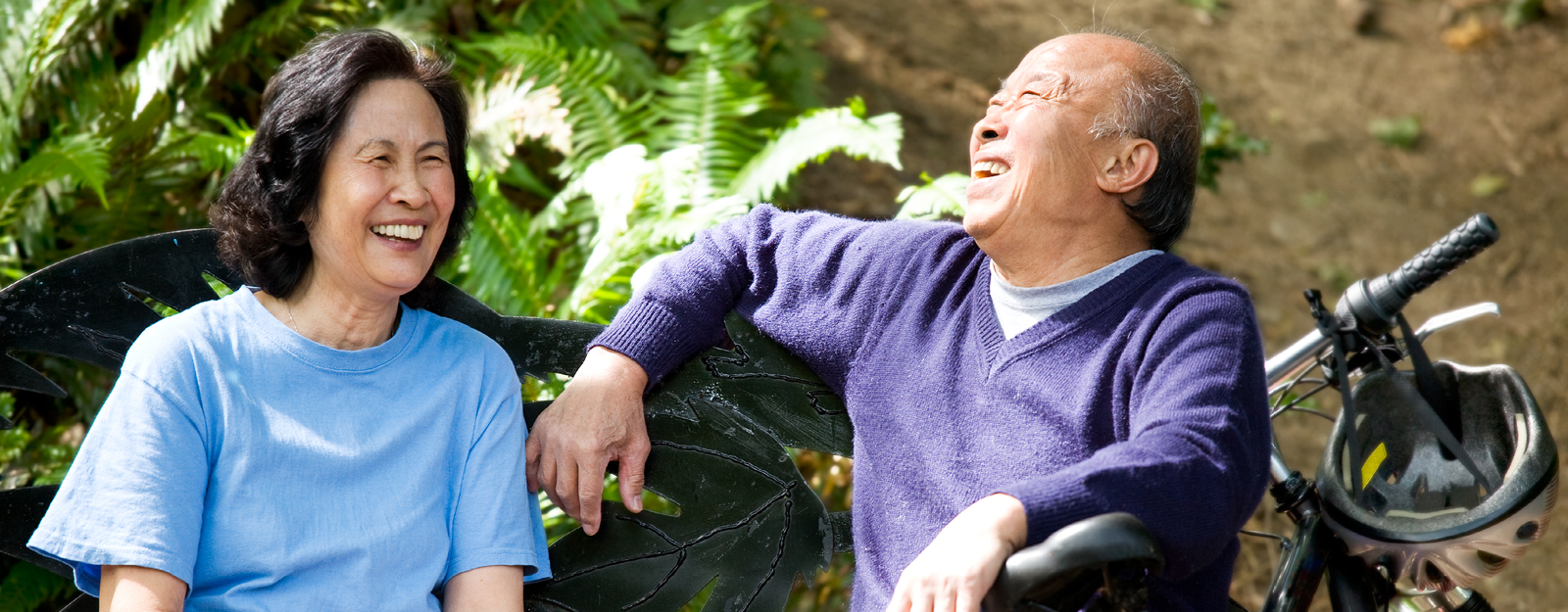 An elderly couple in their 60s sits on a bench and laughs. The man has a mountain bike with a helmet hanging on the handle bars parked beside him.