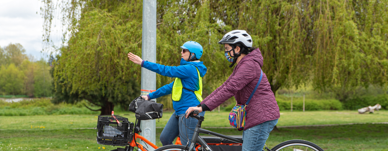 A bike education instructor points at something in the distance. An an adult woman student looks at what the instructor is pointing at. The instructor and student are both on bikes.