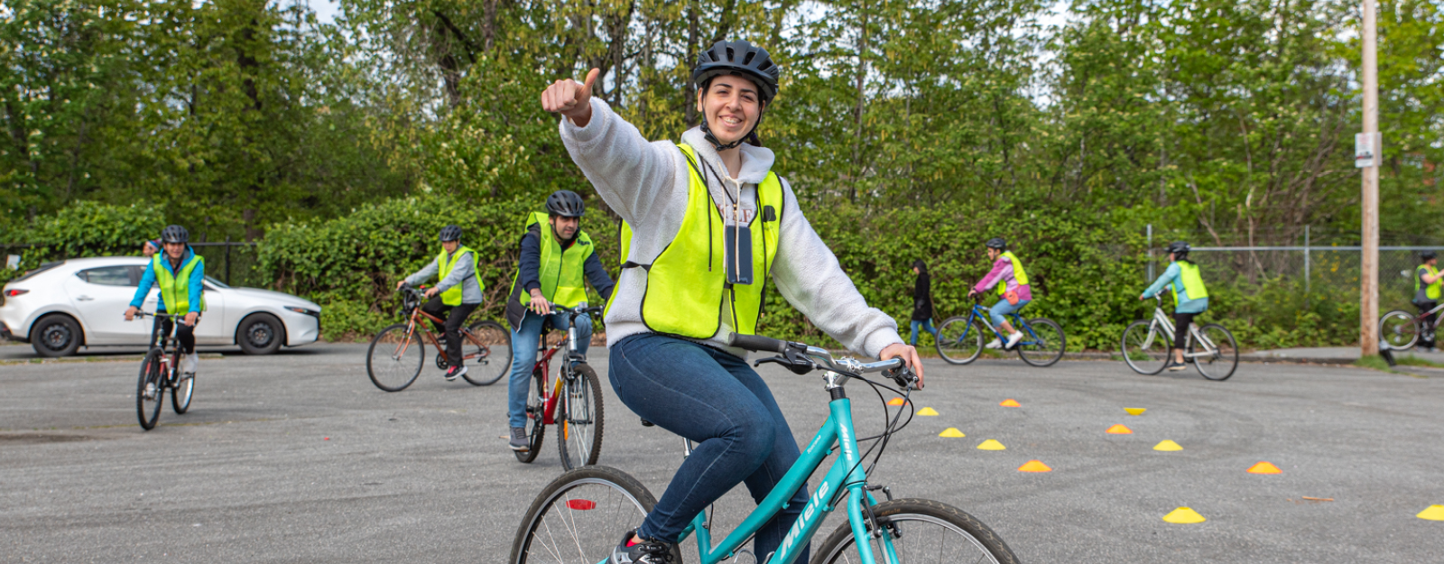 A South Asian adult woman in her 20s rides her bike around a parking lot. She looks at the camera and holds up her thumb in approval. She is smiling. Several other adult students are riding their bikes behind her.