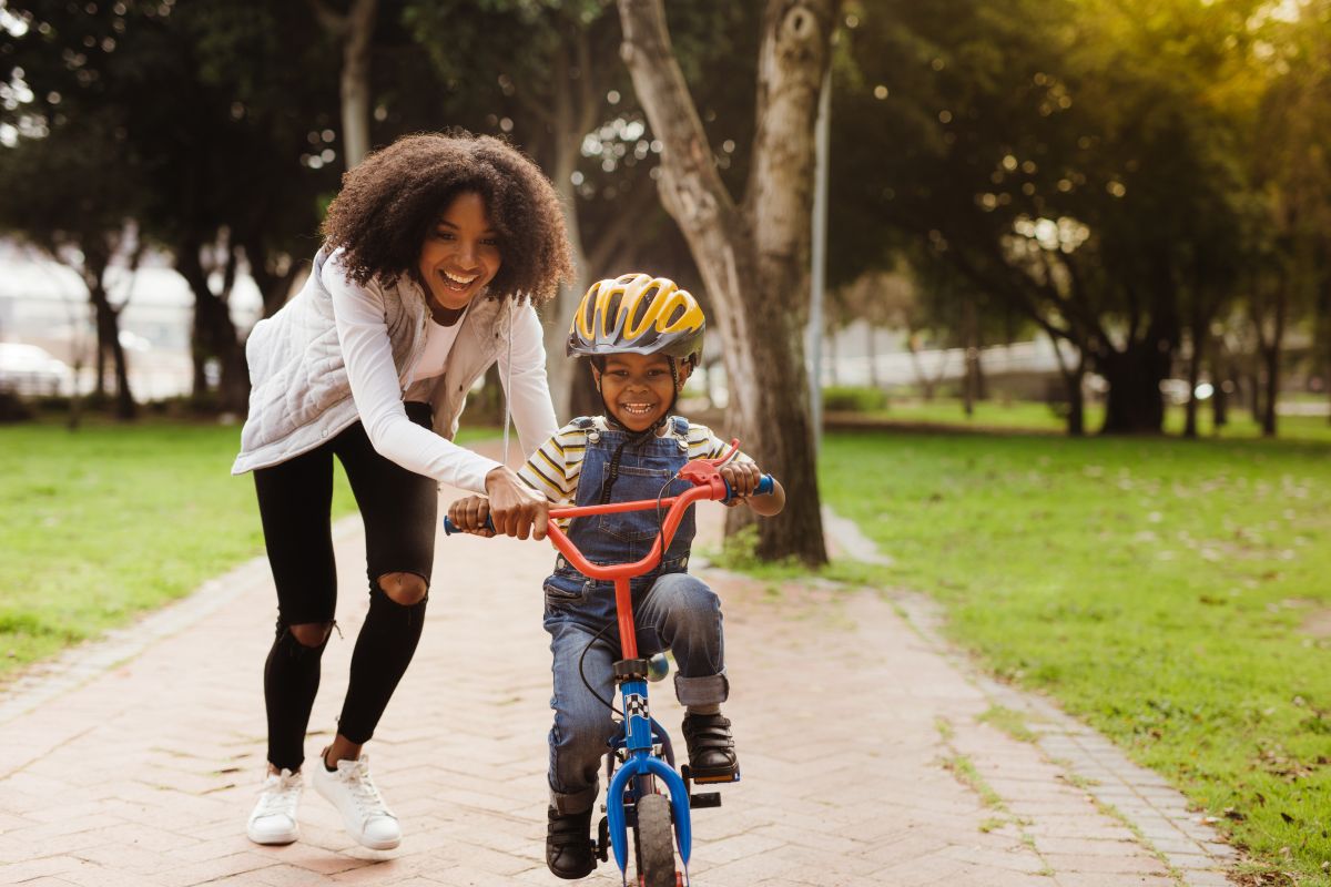 A young mother smiles as she holds on to the back of her young son as he learns to ride his bike. He has a huge smile on his face.