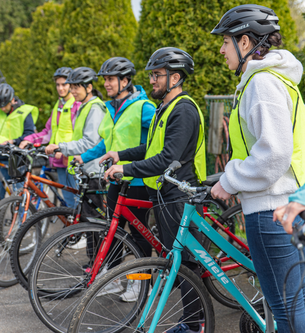Six students stand in a line with their bikes. They are participants of HUB Cycling's Newcomer Bike Mentorship Program. They are each wearing helmets and yellow pinnies.