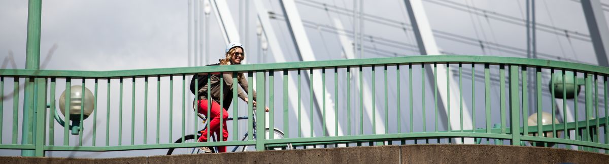 A young woman in her 20s rides her bike across a bridge. She is smiling and having a great time.