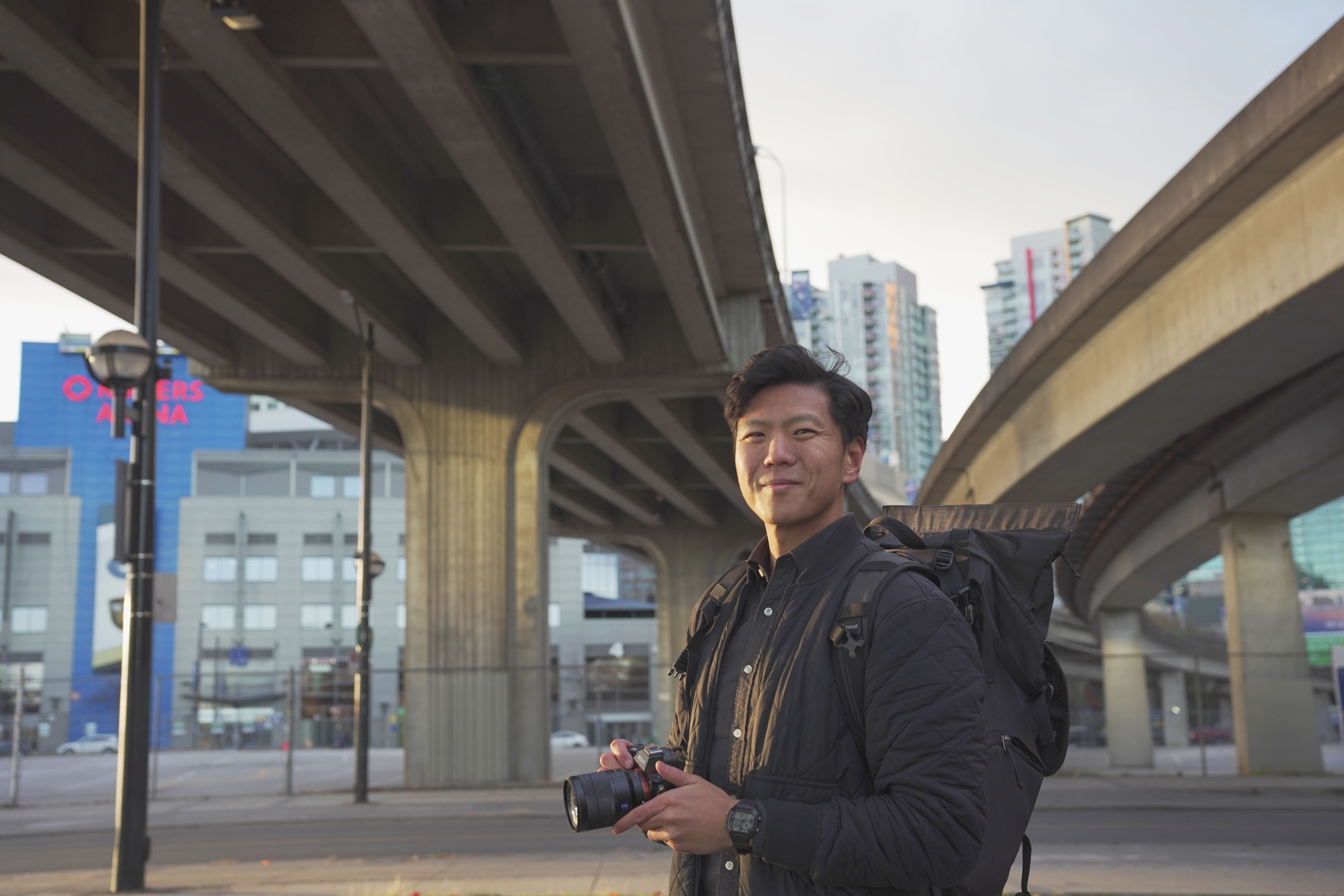 Uytae Lee posing under 2 skytrain tracks holding a camera.