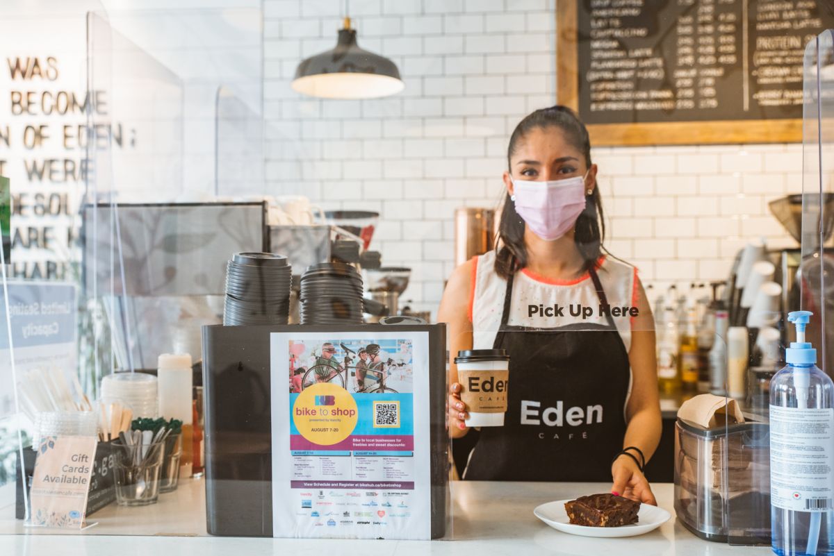 A young South Asian woman in her 20s holds a coffee and baked good behind the counter at Eden Cafe. She is wearing a mask. In the left corner of the photo, we see HUB Cycling's Bike to Shop Poster taped on the divider screen.