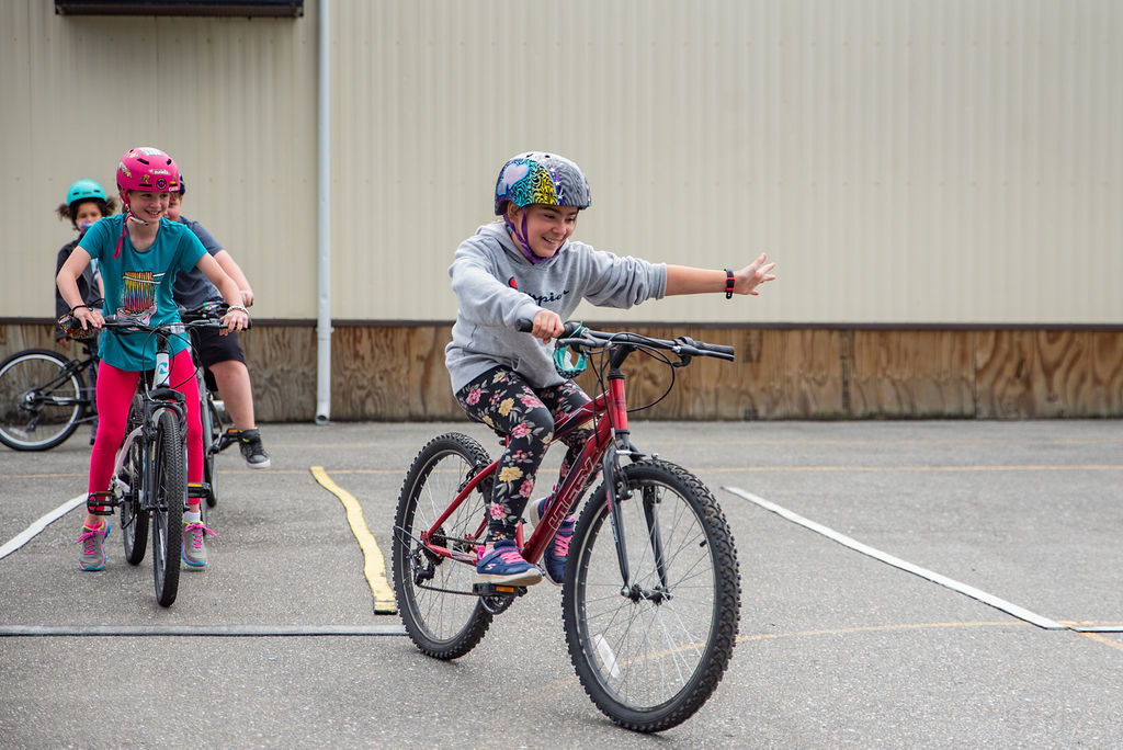 A grade 4/5 female student rides her bike on school grounds. She is smiling and practicing her left-hand turn signal. Several other students follow behind her.