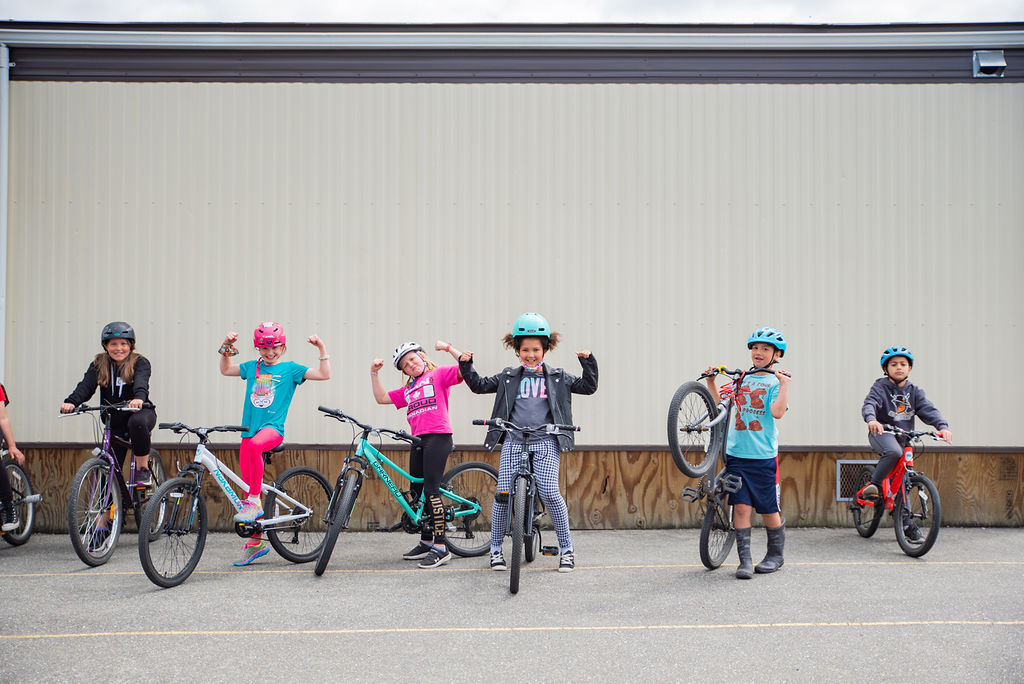 A group of six Grade 4/5 students stand in a row with their bikes on school grounds. They are all smiling for the camera and some students are doing the 'strong-man' pose.