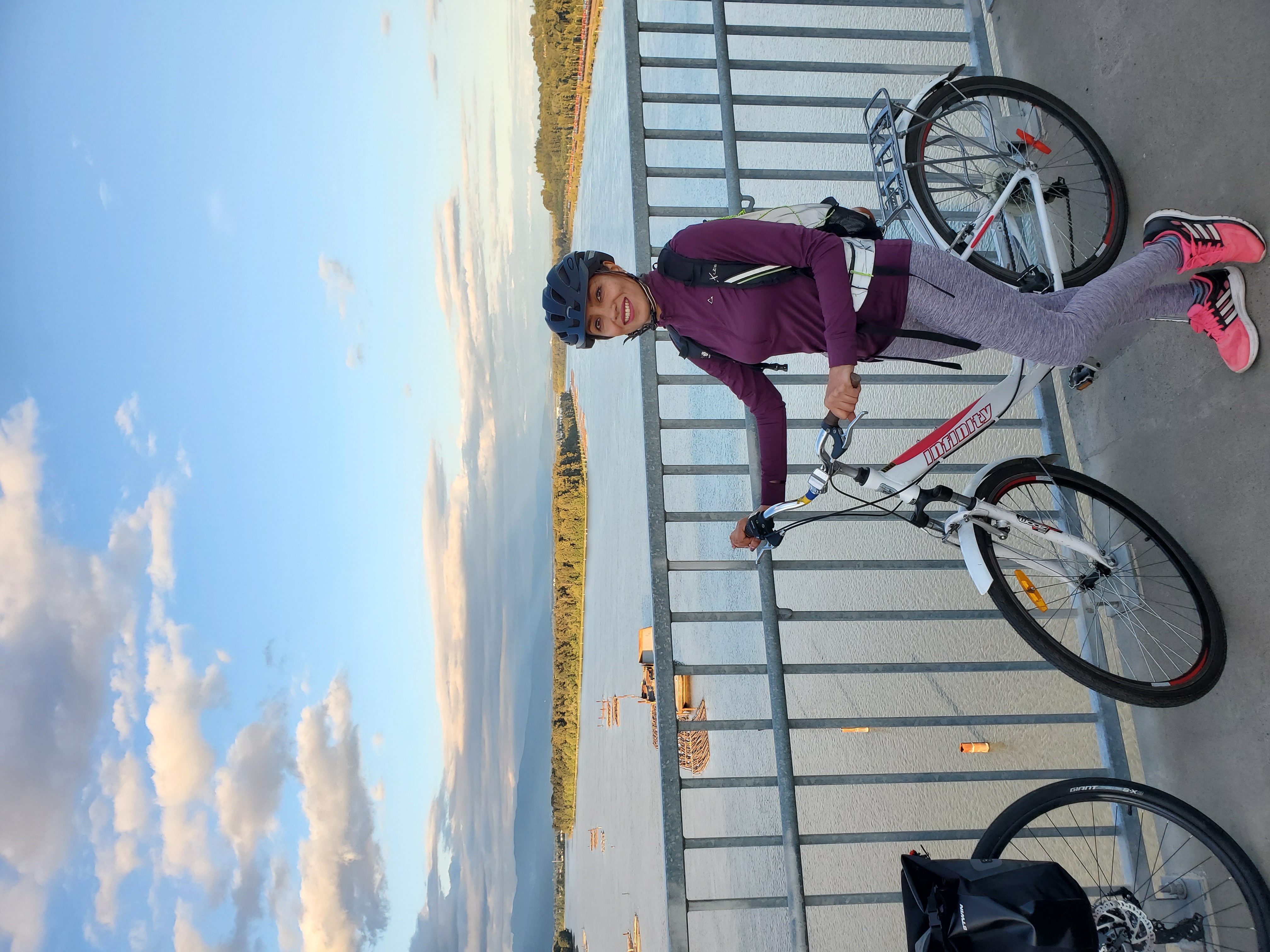 Razieh stands with her bike along the bike path on the Port Mann Bridge. She is wearing gray leggings, running shoes, and a purple long sleeve zip up. We can see the Fraser River below her. The sky is blue with white, fluffy clouds.