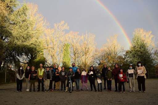 The graduation event from one of Red Fox's Greenest City Biking Programs. About 20 students, HUB Cycling Bike Education Instructors and Red Fox Program Instructors stand in a row on a gravel school field. Everyone is smiling for the camera and some students are holding up graduation certificates. We see a rainbow in the sky behind them.