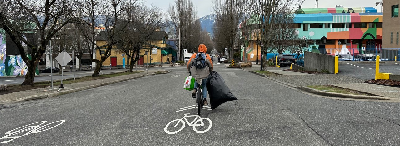 A man cycles away from the camera. He is holding a black garbage bag filled with items and a reusable shopping bag. He is wearing an orange hoodie and a backpack.