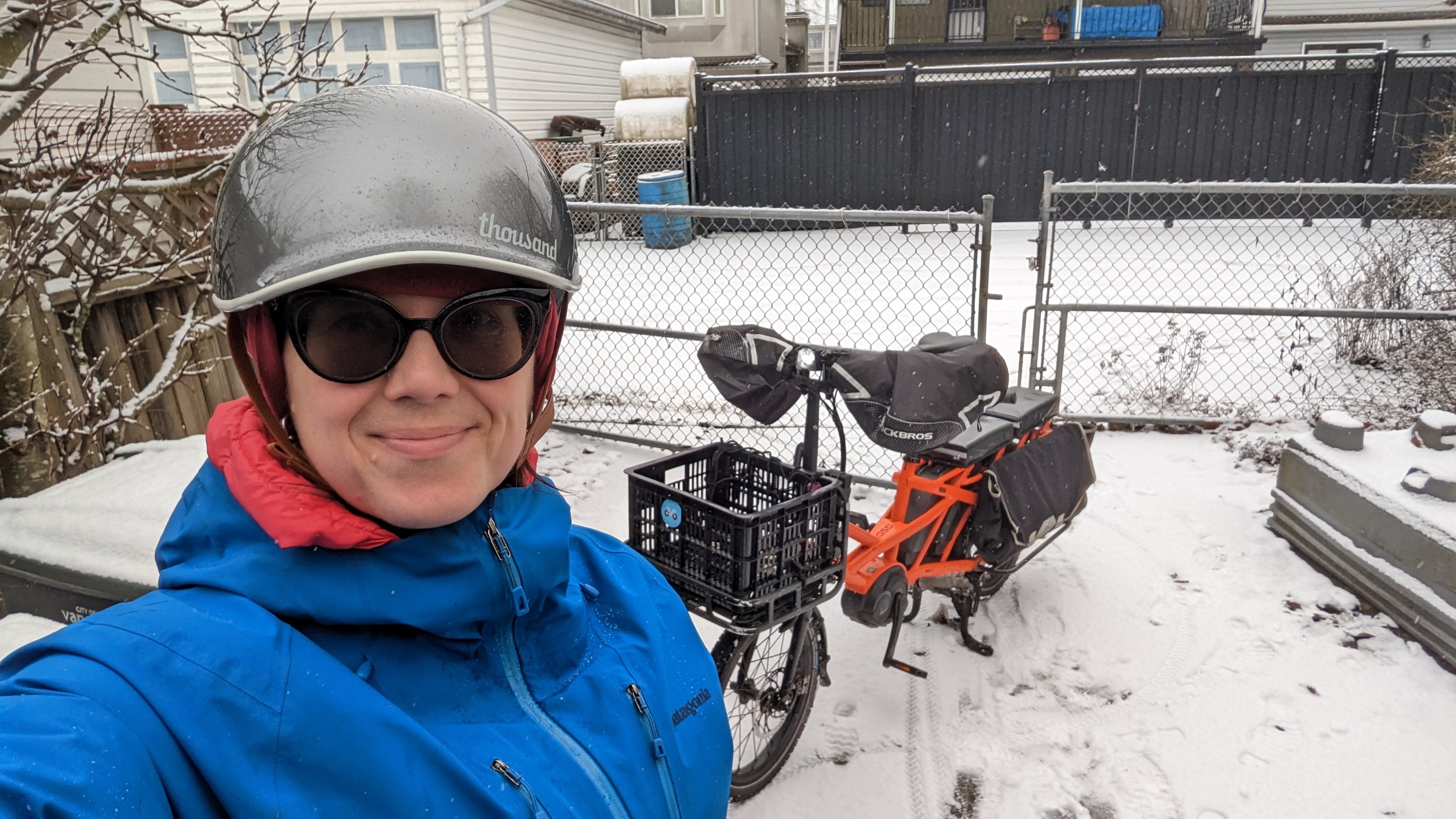 Lisa Corriveau takes a selfie with her electric cargo bike. The ground is covered in snow. She wears a gray helmet, sunglasses, and a blue rain jacket.