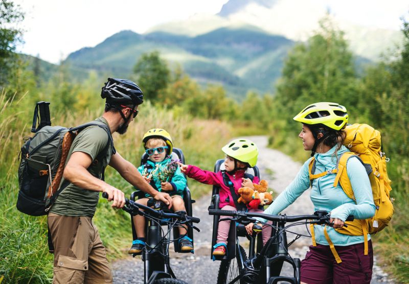 A family including a father, mother and two young children cycle along a gravel trail. We can see the backdrop of a mountain peak behind them.