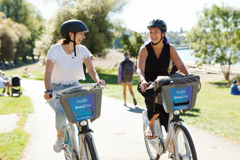 Two women ride Mobi by Shaw Go bikes along the Seawall. They are smiling and laughing. HUB Cycling is partnering with Mobi by Shaw Go to offer free 24 hour passes during Go by Bike Week from May 31 - June 6. The code will be released in mid-May.