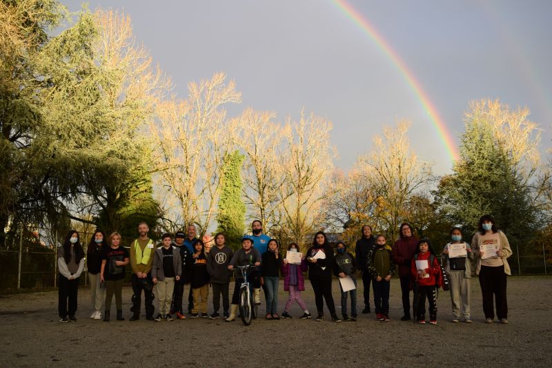 20 students and 4 program coordinators stand in a row outside holding their graduation certificates. There is a rainbow in the sky behind them.