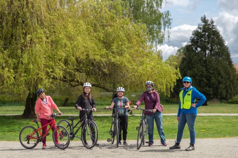 Five women, including four adults participating in a cycling course and one bike education instructor, stand in a field at Trout Lake.