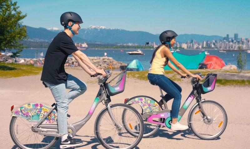 Two young adults ride HUB-branded Mobi by Shaw Go bikes at Jericho Beach.