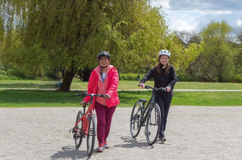 Two adult women wearing helmets walk with their bikes on a gravel field. Both women are smiling.