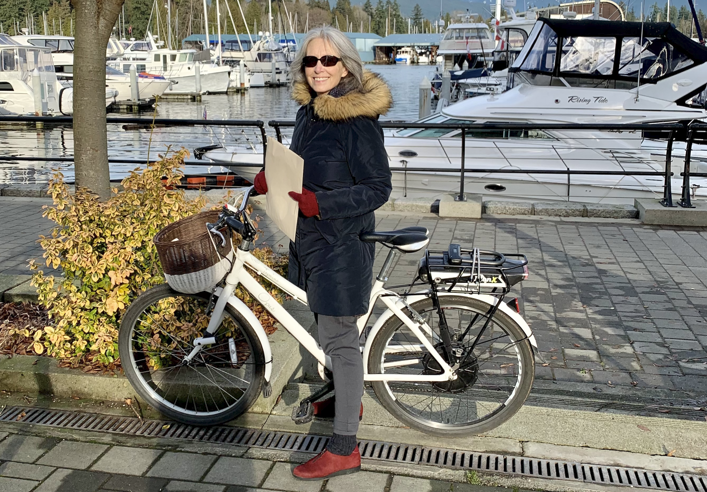 A photo of Dr. Kay Teschke and her bike on the Seawall. We can see the harbor with various boats parked behind her.