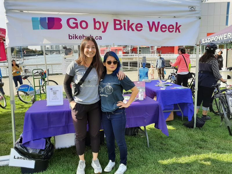 Two women stand and smile for the camera in front of a Go by Bike Week Celebration Station at Science World.